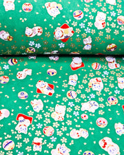 Japanese chiyogami paper featuring assorted white and red Maneki Neko (lucky cats) with small white and gold cherry blossoms and temari balls on a green background with metallic gold details.