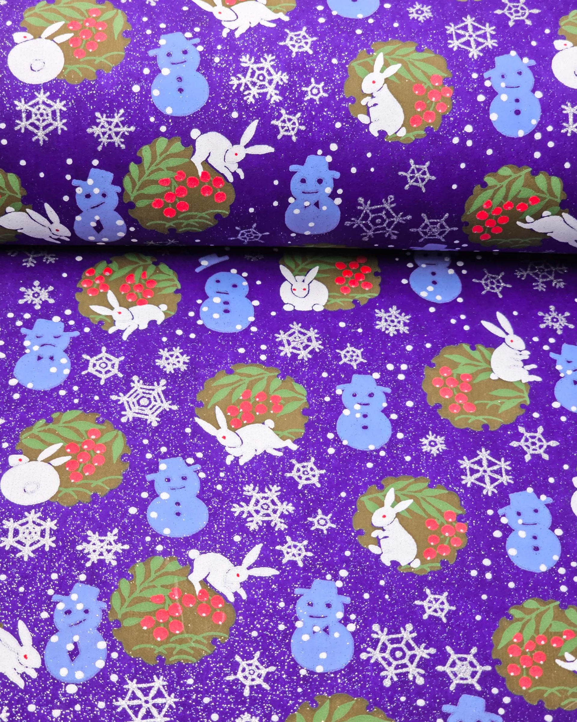 Japanese chiyogami paper featuring light blue snowmen, white rabbits, silver snowflakes, and brown yukiwas with red berries inside on a glitter blue background, shown rolled to highlight the design.
