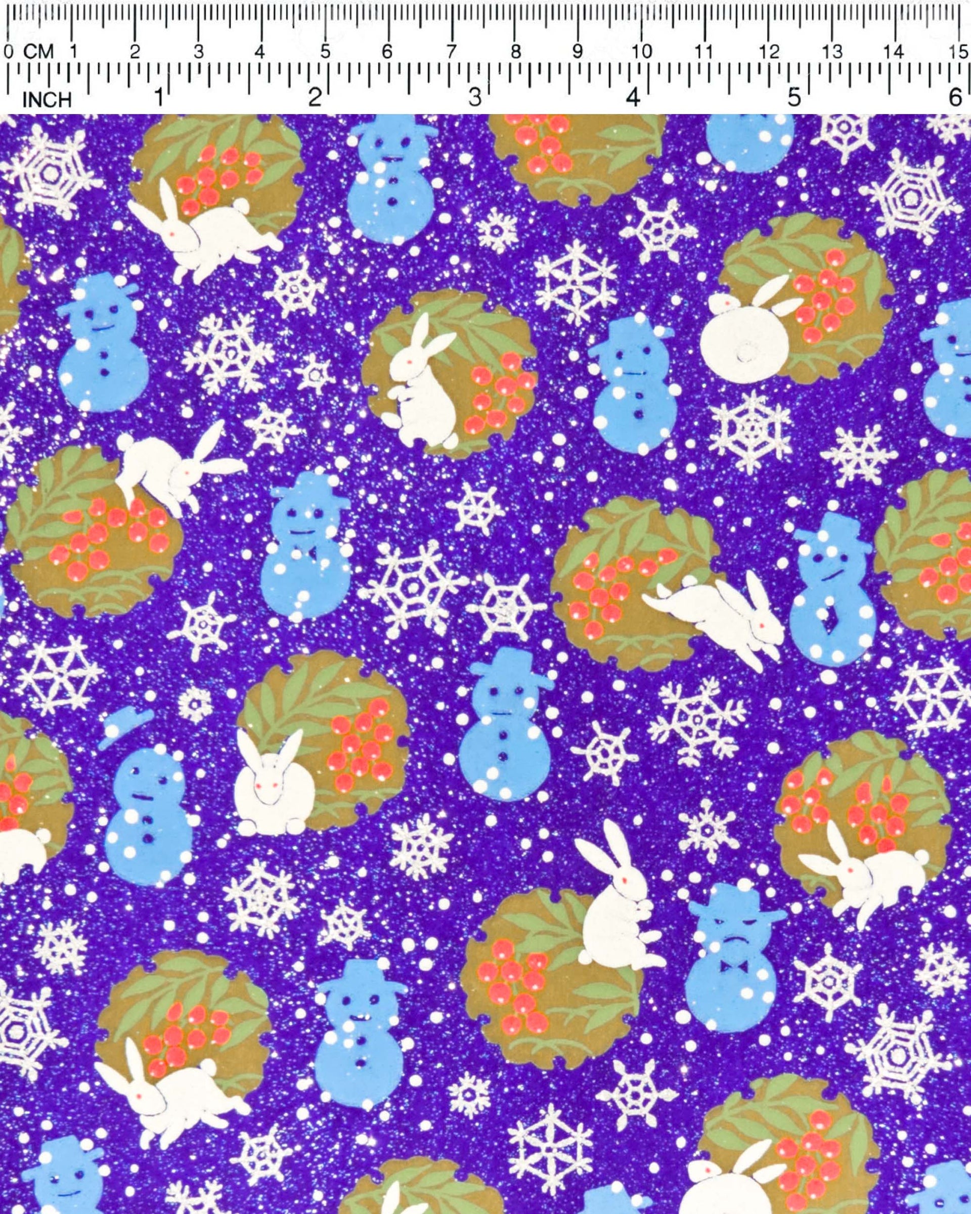 Japanese chiyogami paper featuring light blue snowmen, white rabbits, silver snowflakes, and brown yukiwas with red berries inside on a glitter blue background, shown next to a ruler for size reference.