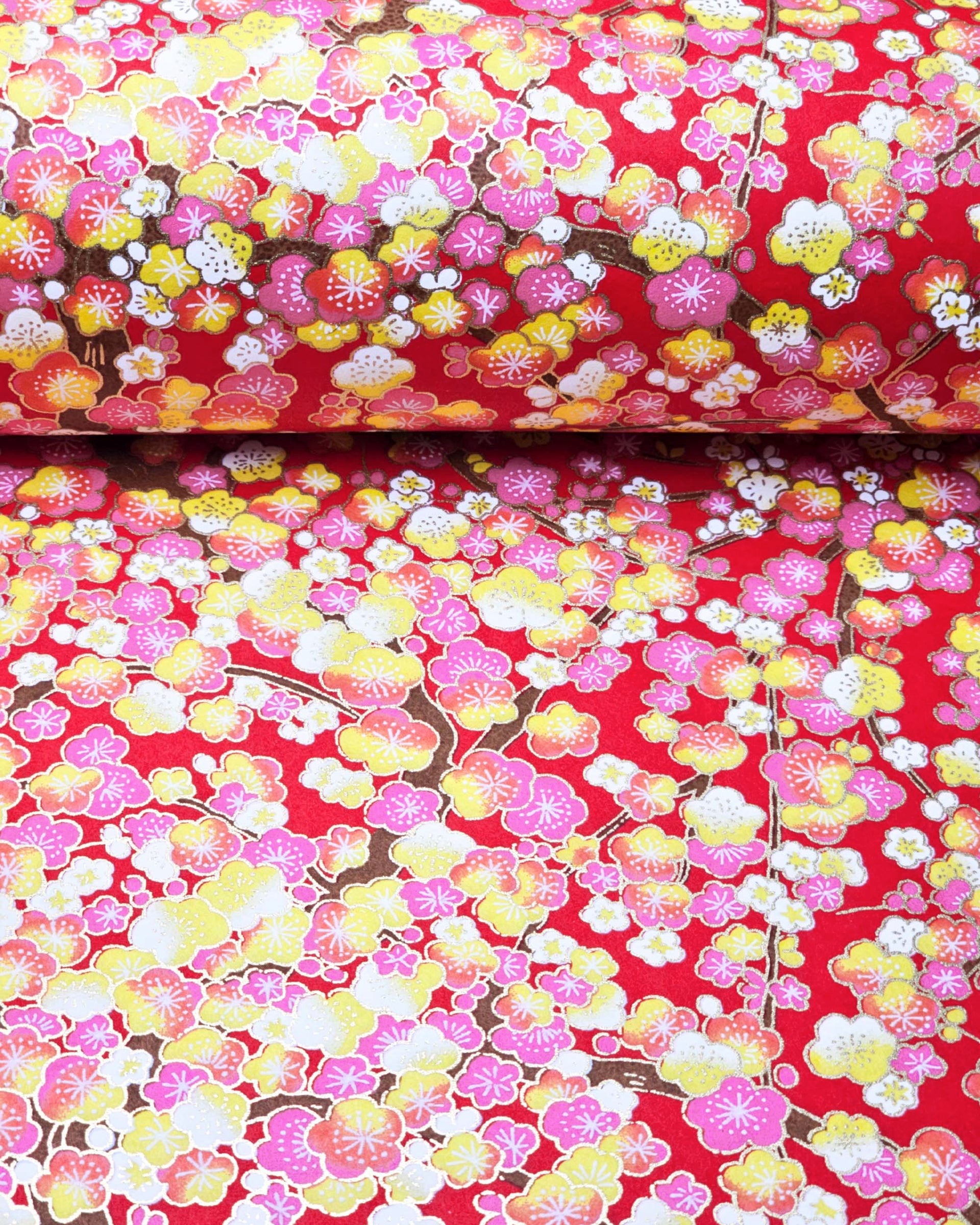 Japanese chiyogami paper featuring pink, yellow, and white plum blossom trees with brown branches on a red background with metallic gold details.