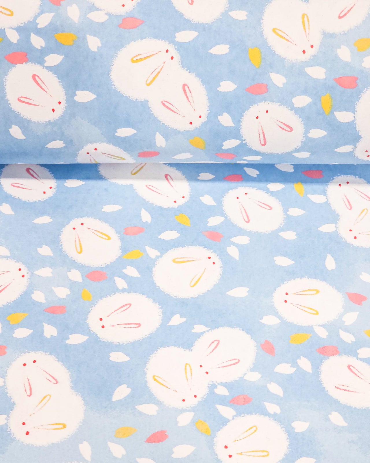 Japanese chiyogami paper featuring round white rabbits with pink and yellow ears surrounded by white, pink, and yellow flower petals on light blue background.