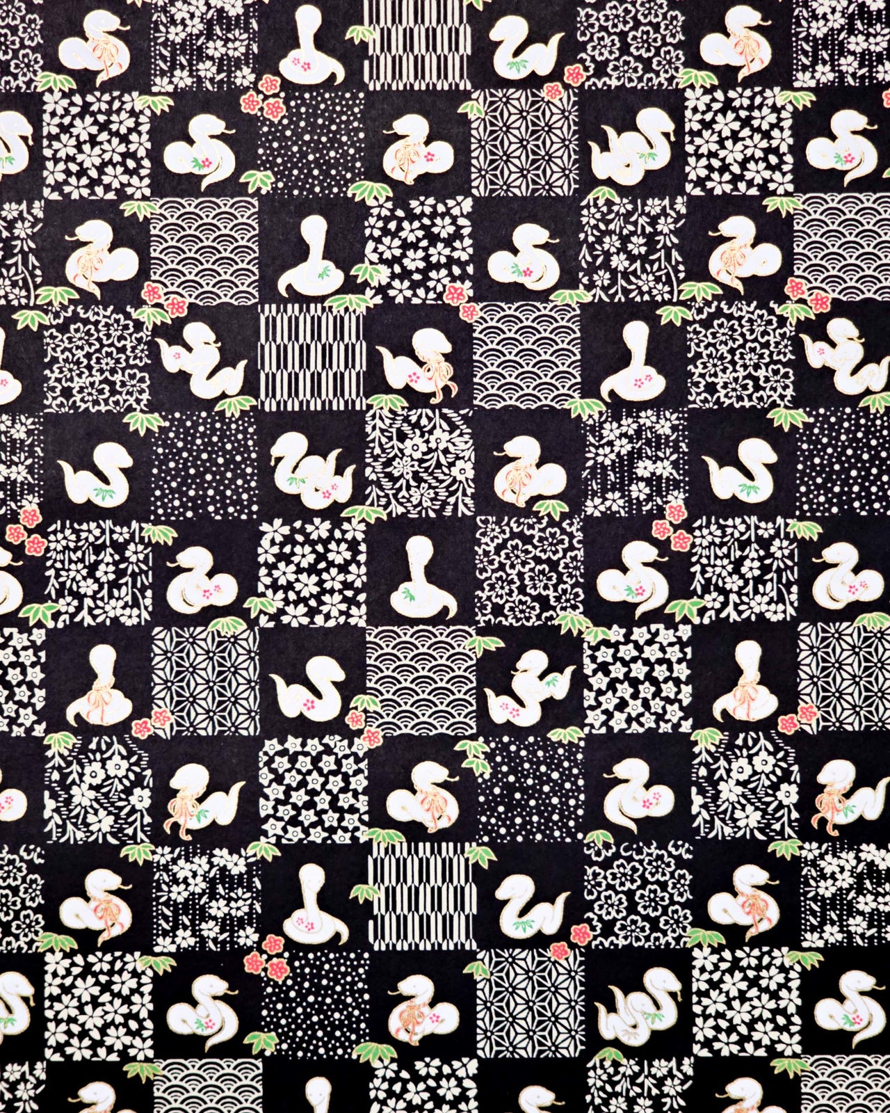 Japanese chiyogami paper featuring white snakes and white assorted traditional Japanese designs in an ichimatsu (checker) design on a black background with metallic gold details. Small red flowers and green leaves accent the design.