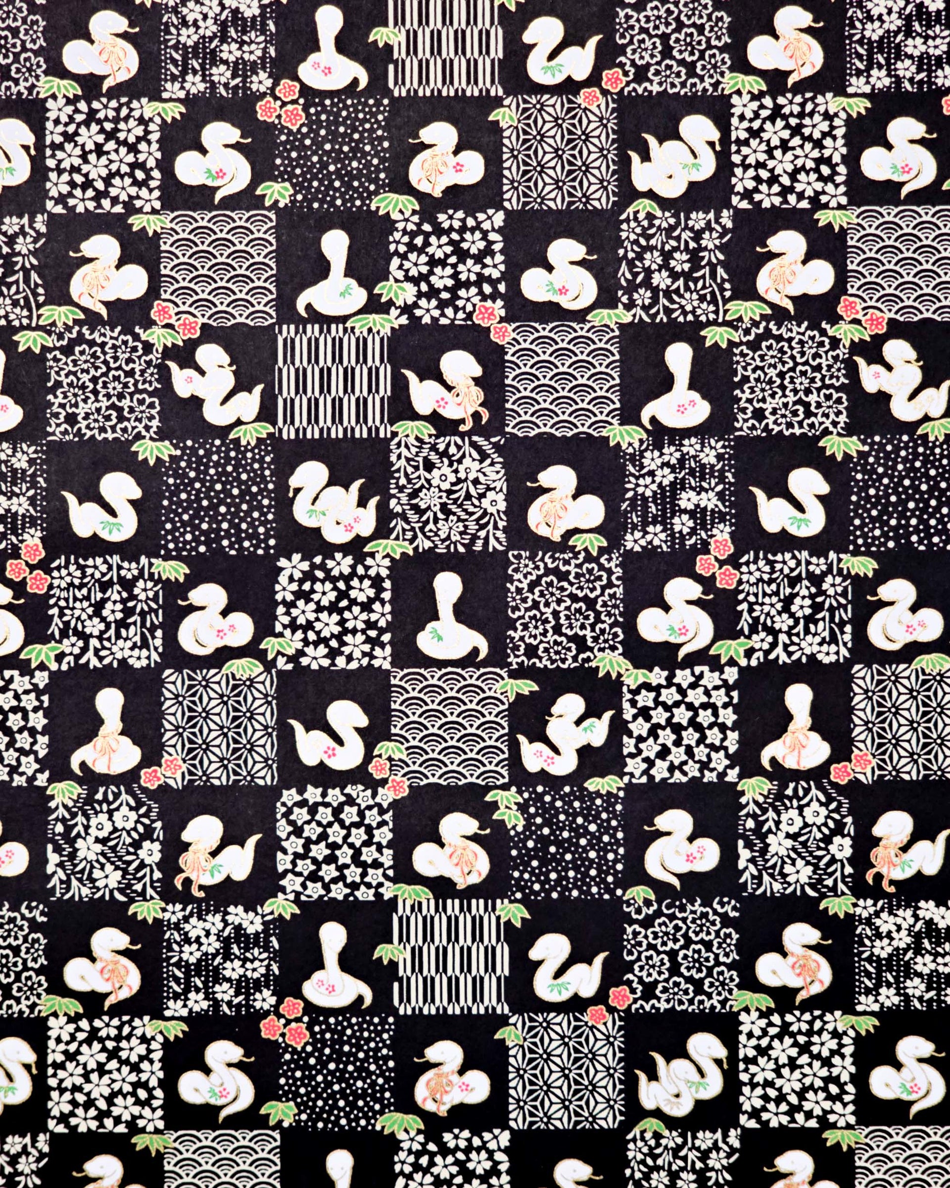 Japanese chiyogami paper featuring white snakes and white assorted traditional Japanese designs in an ichimatsu (checker) design on a black background with metallic gold details. Small red flowers and green leaves accent the design.
