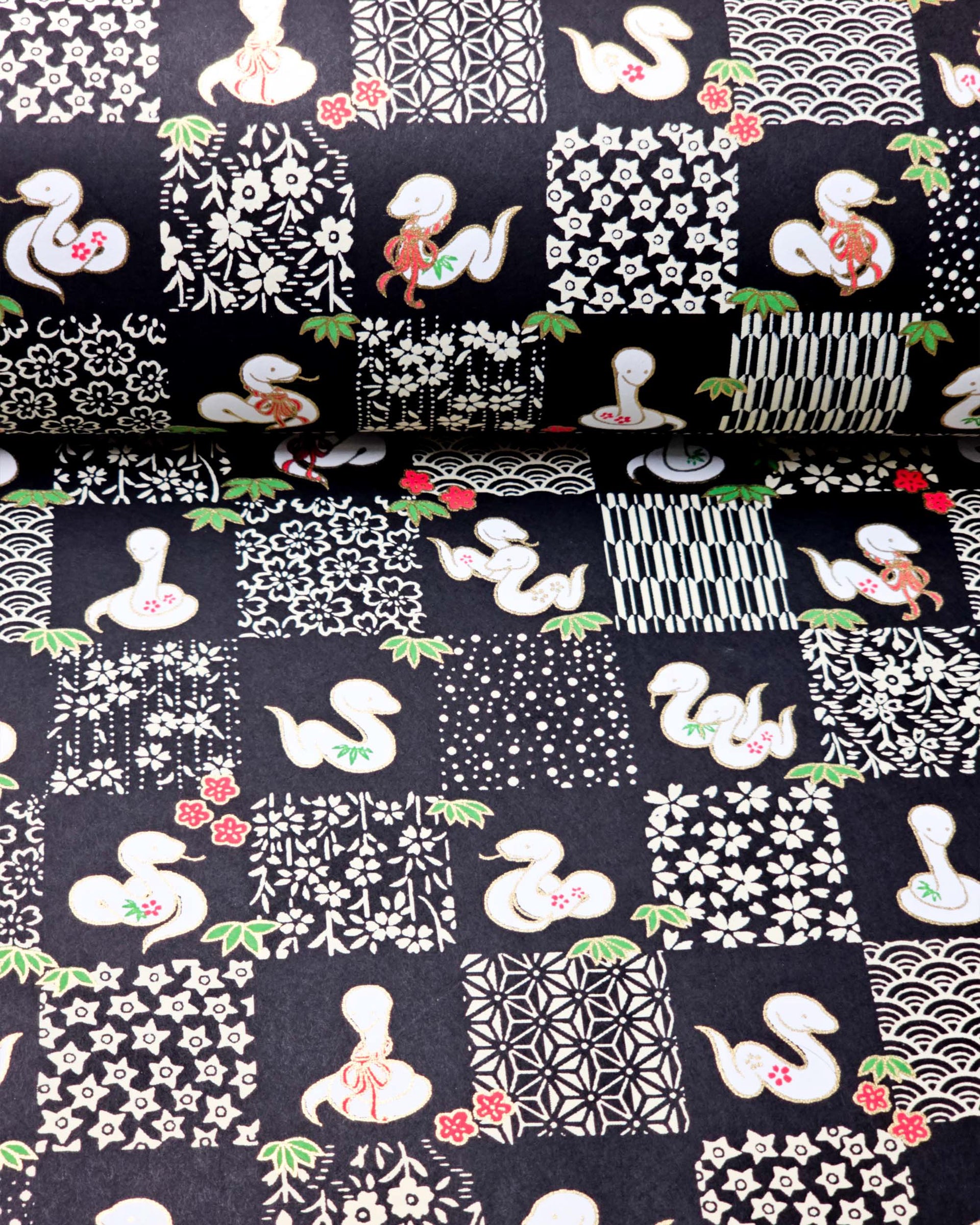 Japanese chiyogami paper featuring white snakes and white assorted traditional Japanese designs in an ichimatsu (checker) design on a black background with metallic gold details. Small red flowers and green leaves accent the design.