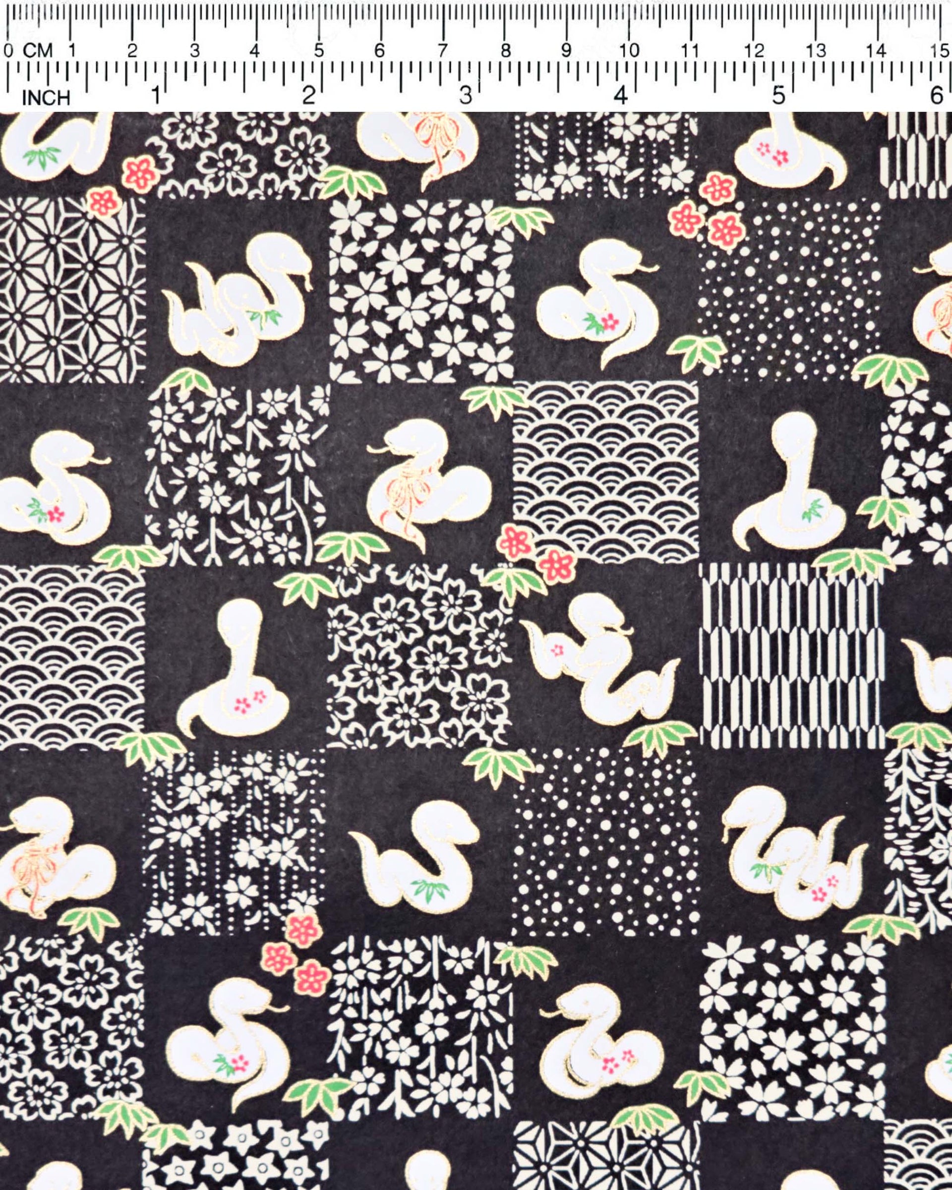 Japanese chiyogami paper featuring white snakes and white assorted traditional Japanese designs in an ichimatsu (checker) design on a black background with metallic gold details. Small red flowers and green leaves accent the design.