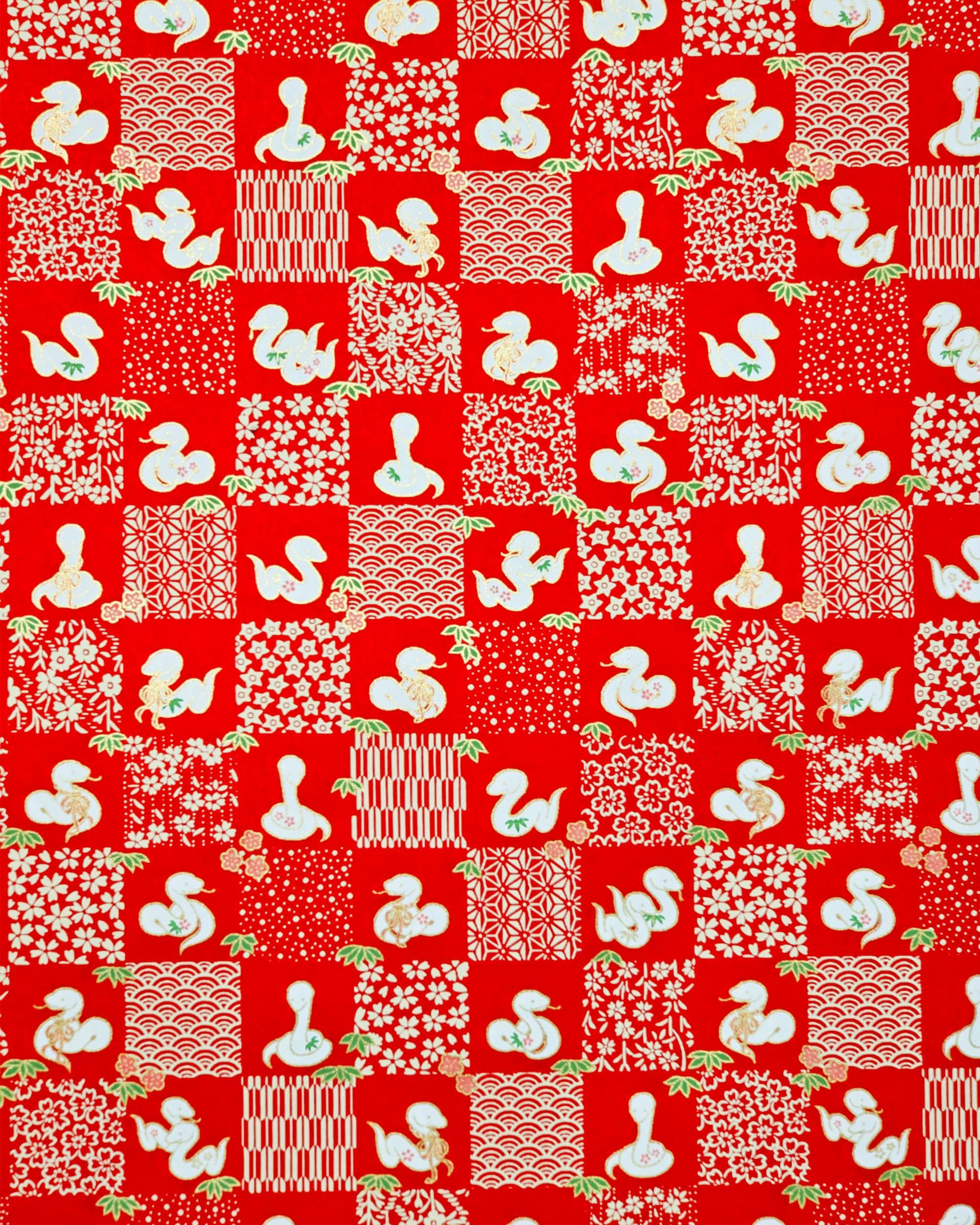 Japanese chiyogami paper featuring white snakes and white assorted traditional Japanese designs in an ichimatsu (checker) design on a red background with metallic gold details. Small pink flowers and green leaves accent the design.