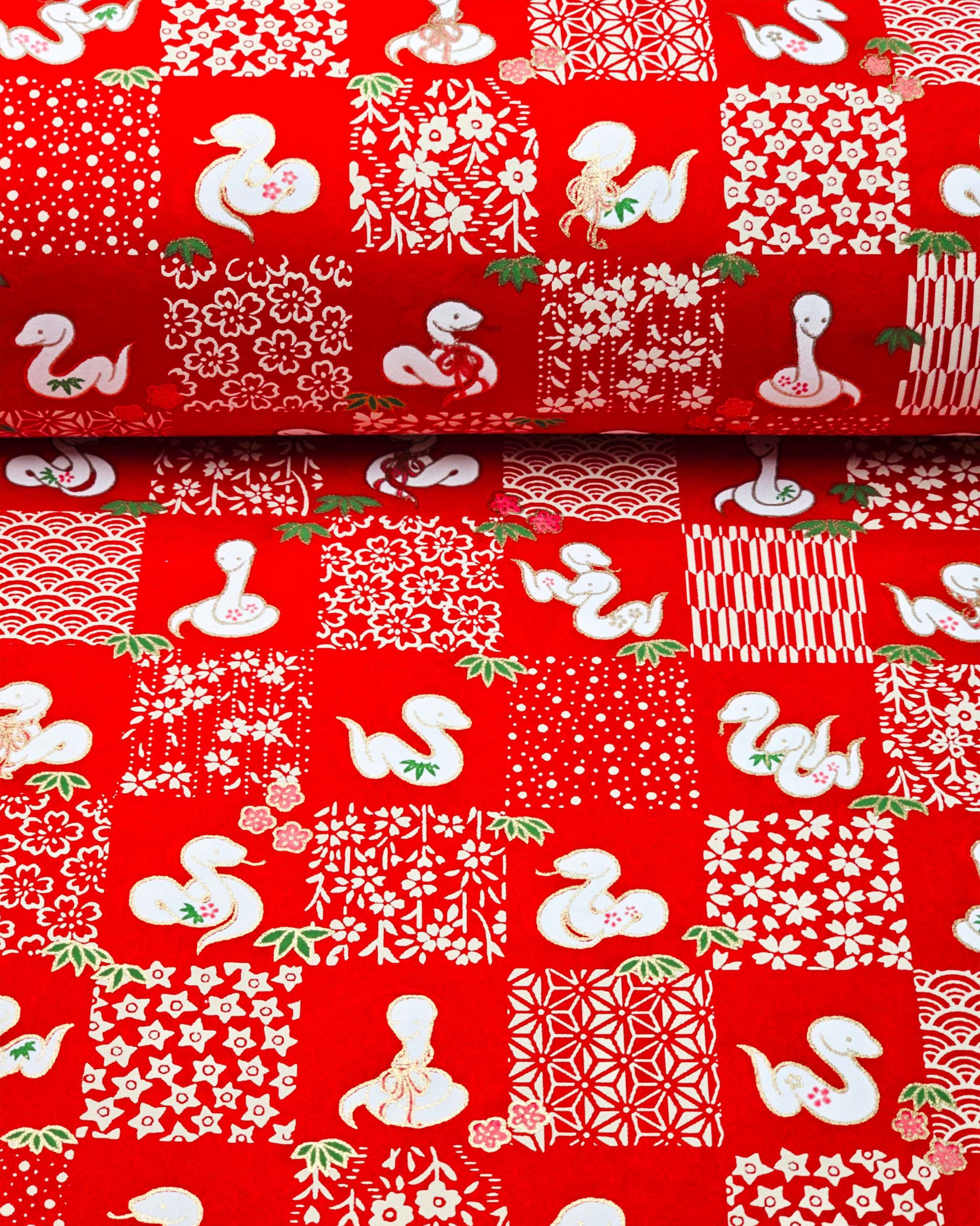 Japanese chiyogami paper featuring white snakes and white assorted traditional Japanese designs in an ichimatsu (checker) design on a red background with metallic gold details. Small pink flowers and green leaves accent the design.