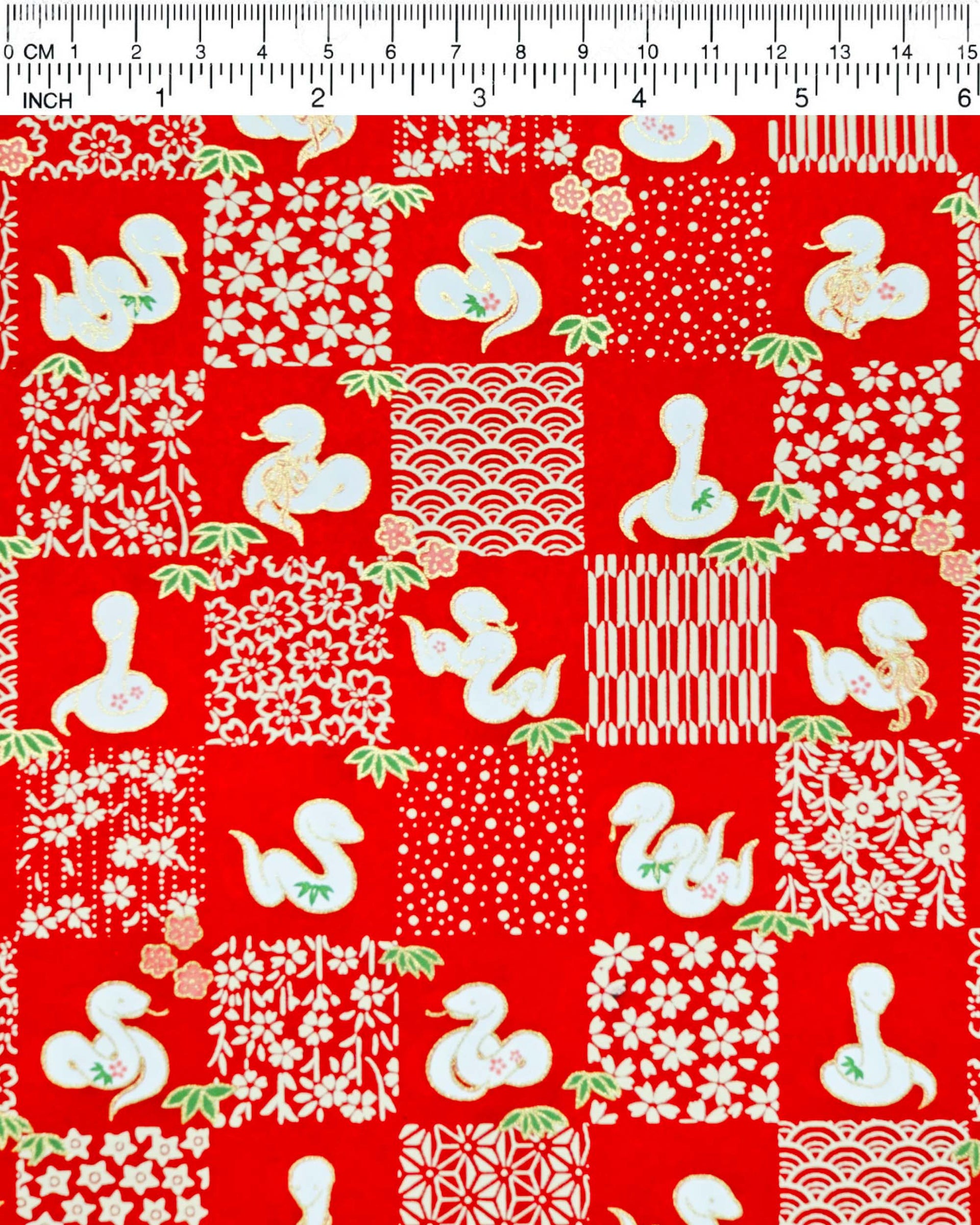 Japanese chiyogami paper featuring white snakes and white assorted traditional Japanese designs in an ichimatsu (checker) design on a red background with metallic gold details. Small pink flowers and green leaves accent the design.