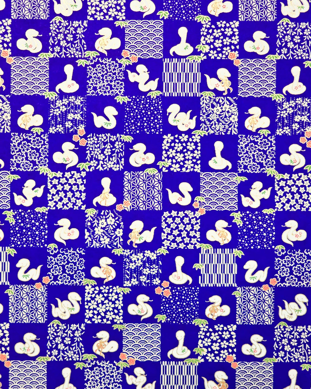 Japanese chiyogami paper featuring white snakes and white assorted traditional Japanese designs in an ichimatsu (checker) design on a blue background with metallic gold details. Small red flowers and green leaves accent the design.