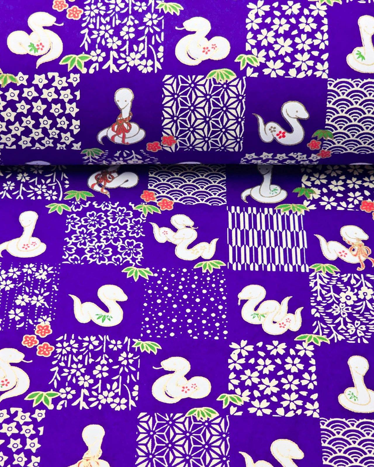 Japanese chiyogami paper featuring white snakes and white assorted traditional Japanese designs in an ichimatsu (checker) design on a blue background with metallic gold details. Small red flowers and green leaves accent the design.