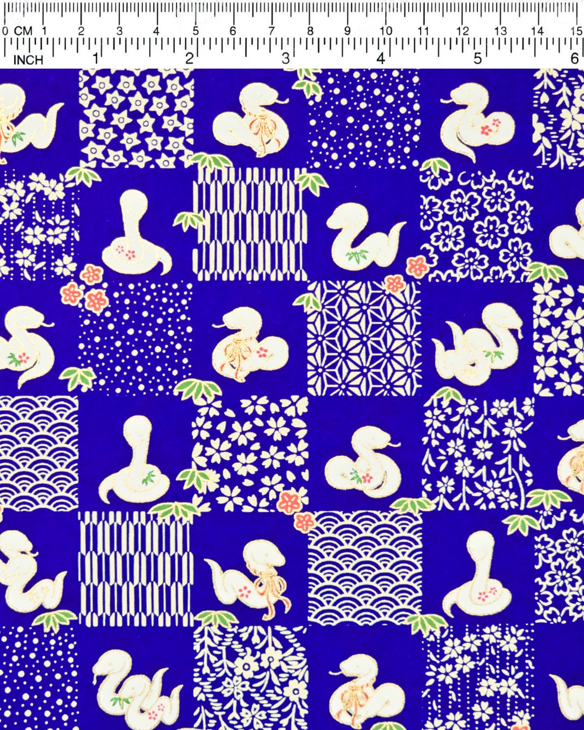 Japanese chiyogami paper featuring white snakes and white assorted traditional Japanese designs in an ichimatsu (checker) design on a blue background with metallic gold details. Small red flowers and green leaves accent the design.