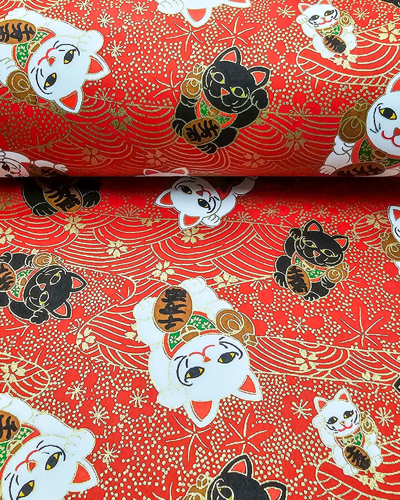 Japanese chiyogami paper featuring large black and white maneki neko (lucky cats) on a red background with metallic gold details. The red background has metallic gold cherry blossoms, plum blossoms, maples leaves, and wave crests.