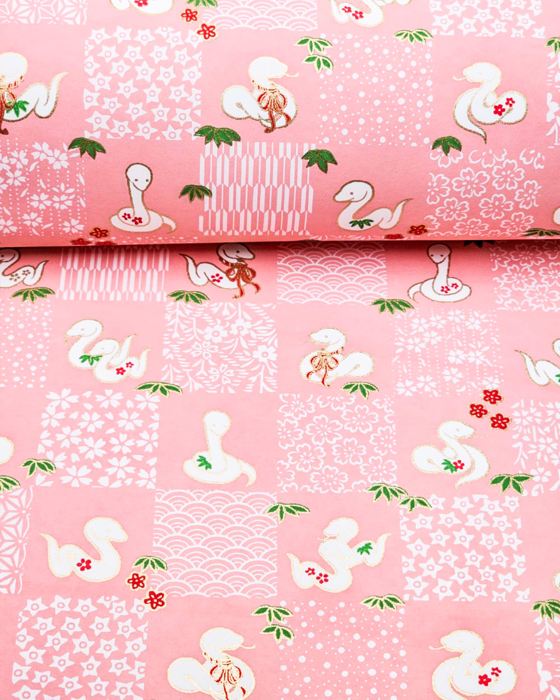 Japanese chiyogami paper featuring white snakes and white assorted traditional Japanese designs in an ichimatsu (checker) design on a pink background with metallic gold details.