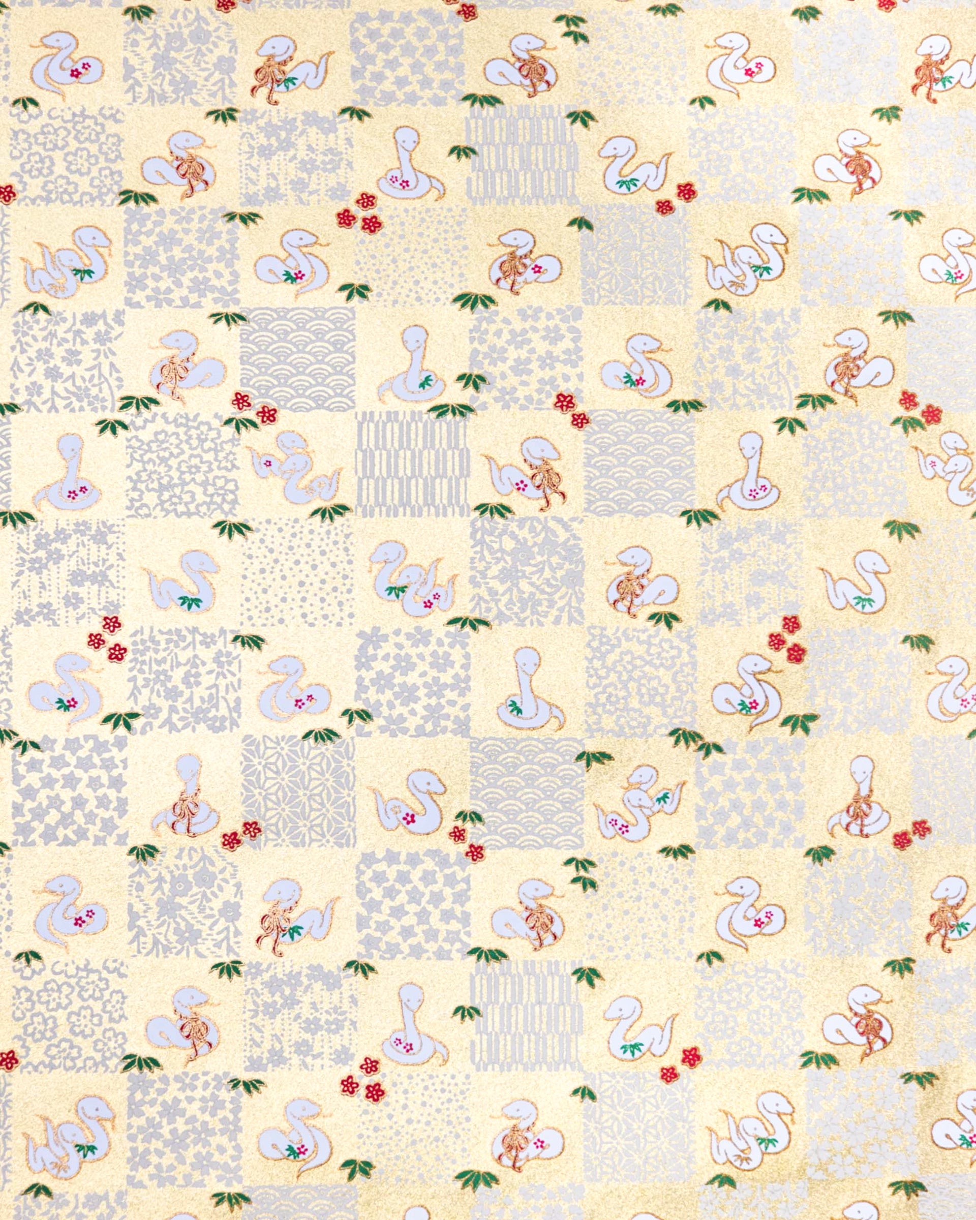 Japanese chiyogami paper featuring white snakes and white assorted traditional Japanese designs in an ichimatsu (checker) design on a gold background with metallic gold details. Small red plum blossoms and green bamboo leaves are scattered throughout the design.