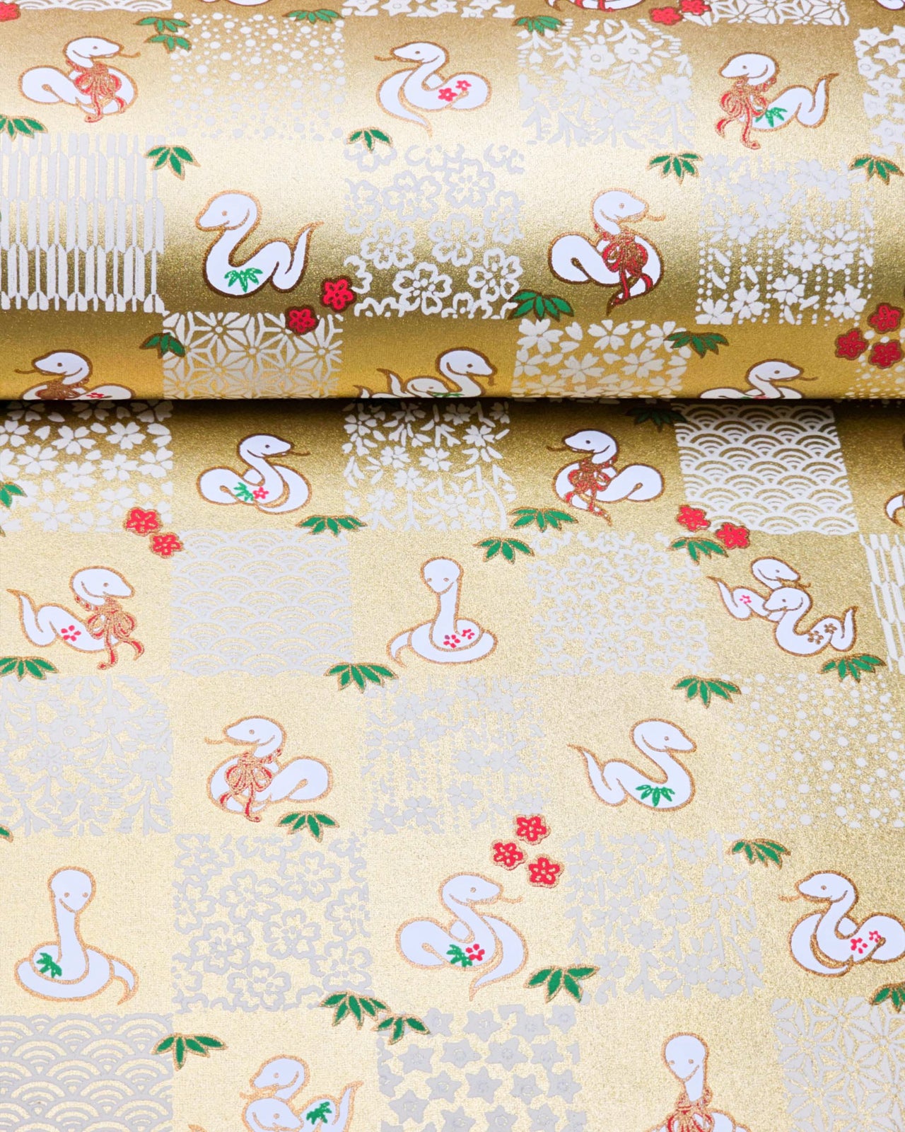 Japanese chiyogami paper featuring white snakes and white assorted traditional Japanese designs in an ichimatsu (checker) design on a gold background with metallic gold details. Small red plum blossoms and green bamboo leaves are scattered throughout the design.