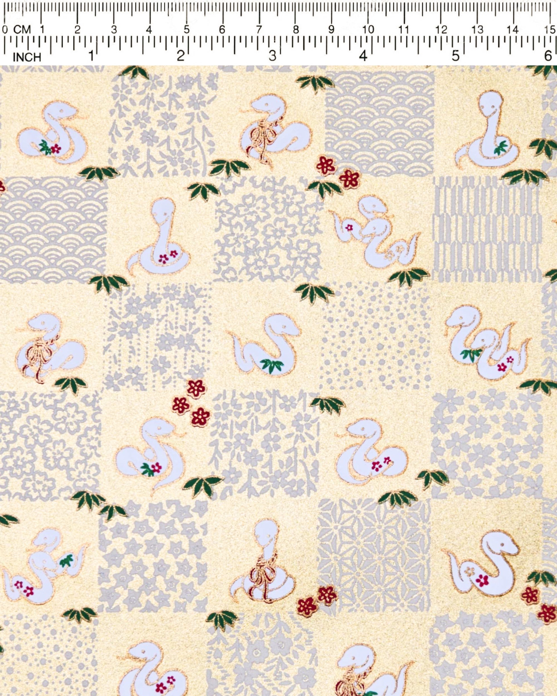Japanese chiyogami paper featuring white snakes and white assorted traditional Japanese designs in an ichimatsu (checker) design on a gold background with metallic gold details. Small red plum blossoms and green bamboo leaves are scattered throughout the design.
