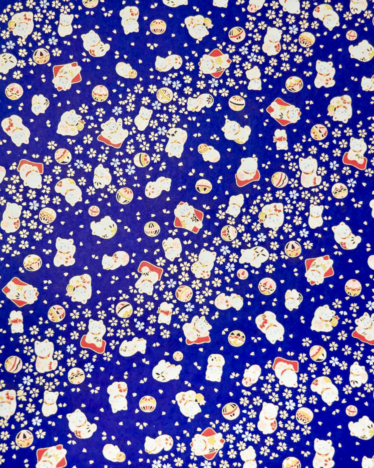 Japanese chiyogami paper featuring white and red Maneki Neko (lucky cats) with small white and gold cherry blossoms and temari balls on a dark blue background with metallic gold details.