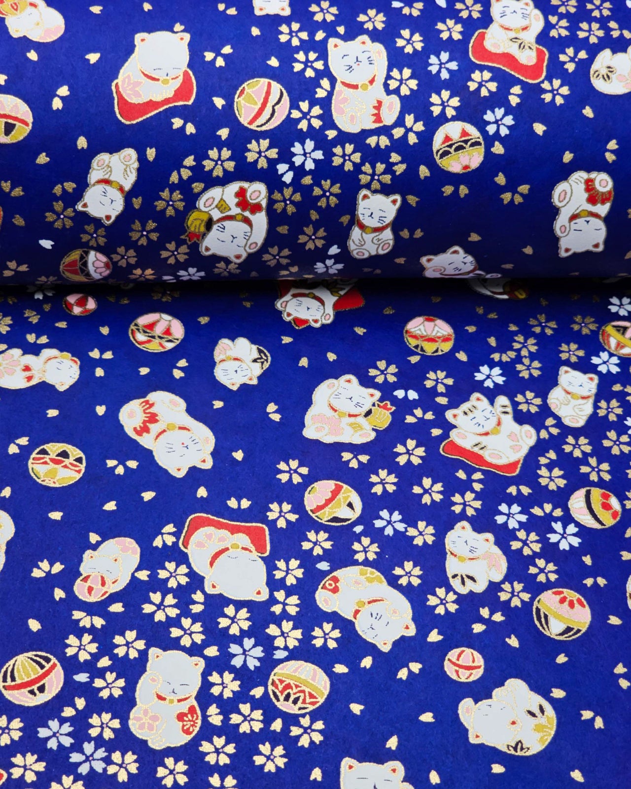 Japanese chiyogami paper featuring white and red Maneki Neko (lucky cats) with small white and gold cherry blossoms and temari balls on a dark blue background with metallic gold details.