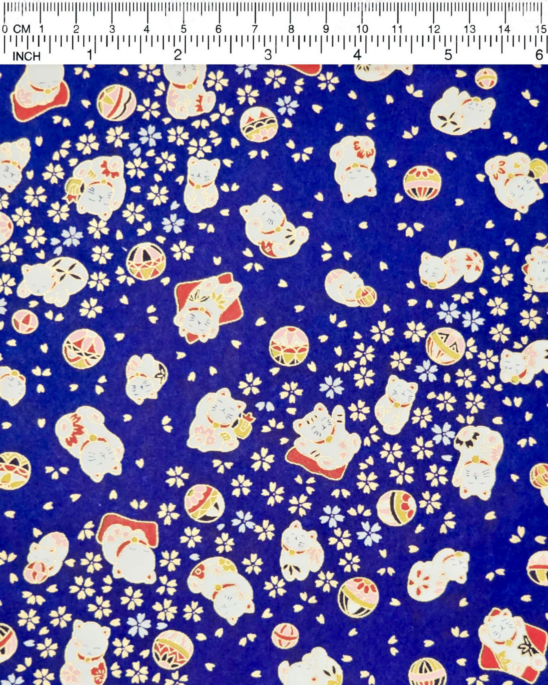 Japanese chiyogami paper featuring white and red Maneki Neko (lucky cats) with small white and gold cherry blossoms and temari balls on a dark blue background with metallic gold details.