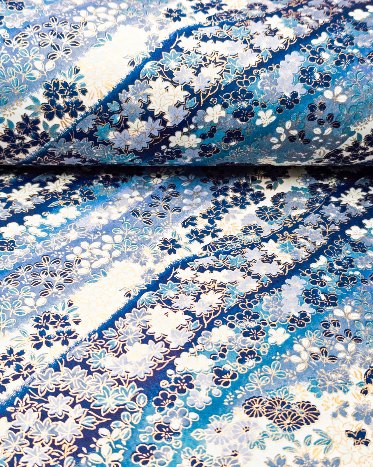 Japanese chiyogami paper featuring an assortment of blue and white traditional Japanese flowers and leaves on a blue and white gradient background with metallic gold details, shown rolled to highlight the design.