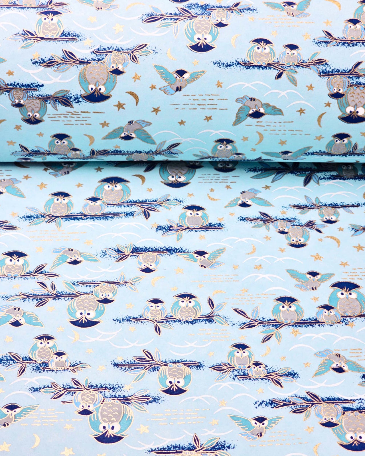 Japanese chiyogami paper featuring blue and gray owls perched on branches and flying around with a light blue night sky background adorned with small metallic gold stars and crescent moons, shown rolled to highlight the design.
