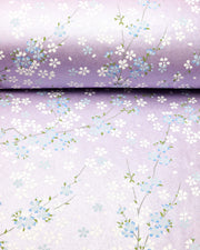 Japanese chiyogami paper featuring light blue and white cherry blossoms with green leaves and branches on a pearlescent light purple background.