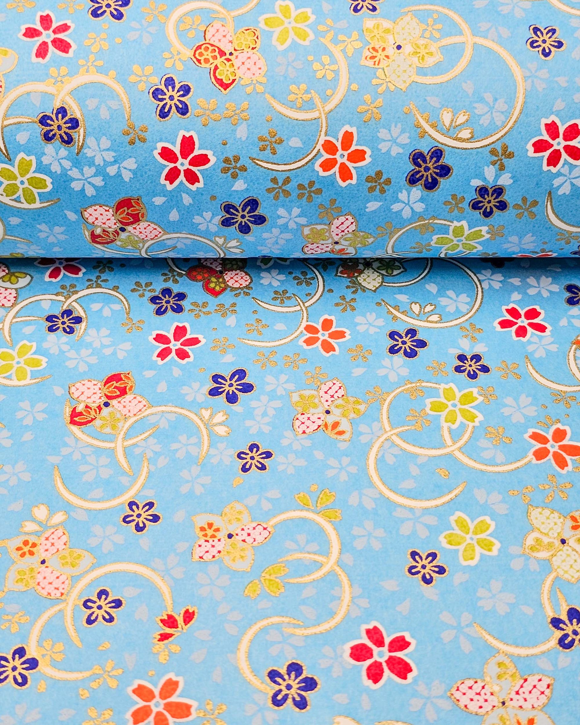 Japanese chiyogami paper featuring red, blue, green, and white plum and cherry blossoms on a sky blue background with metallic gold accents.