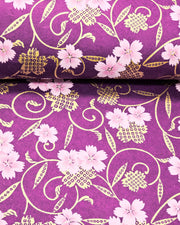 Japanese chiyogami paper featuring pink cherry blossoms alongside gold cherry blossoms and leaves decorated with a traditional kanoko (fawn) pattern on a purple background with metallic gold details, shown rolled to highlight the design.
