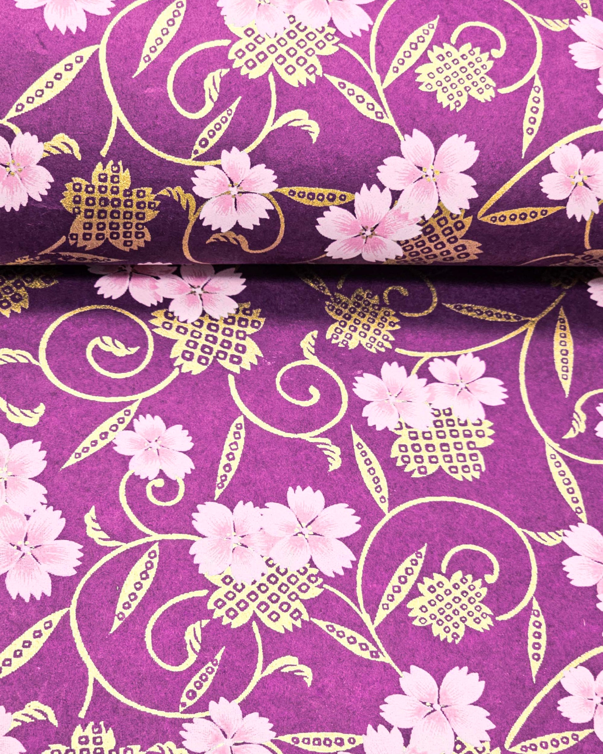 Japanese chiyogami paper featuring pink cherry blossoms alongside gold cherry blossoms and leaves decorated with a traditional kanoko (fawn) pattern on a purple background with metallic gold details, shown rolled to highlight the design.
