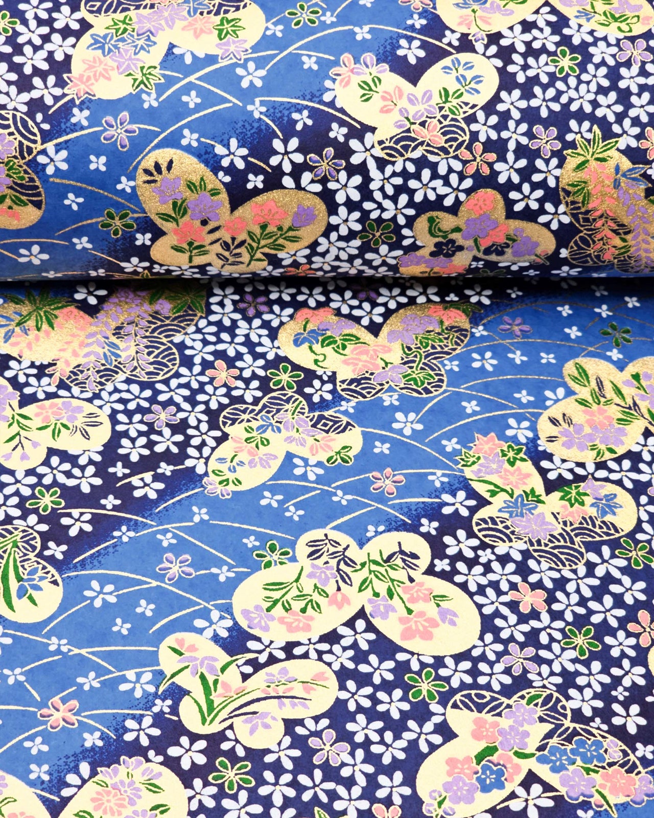 Japanese chiyogami paper featuring metallic gold butterflies with purple and pink flowers on a blue background covered with small white flowers, shown rolled to highlight the design.