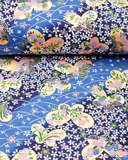 Japanese chiyogami paper featuring metallic gold butterflies with purple and pink flowers on a blue background covered with small white flowers, shown rolled to highlight the design.