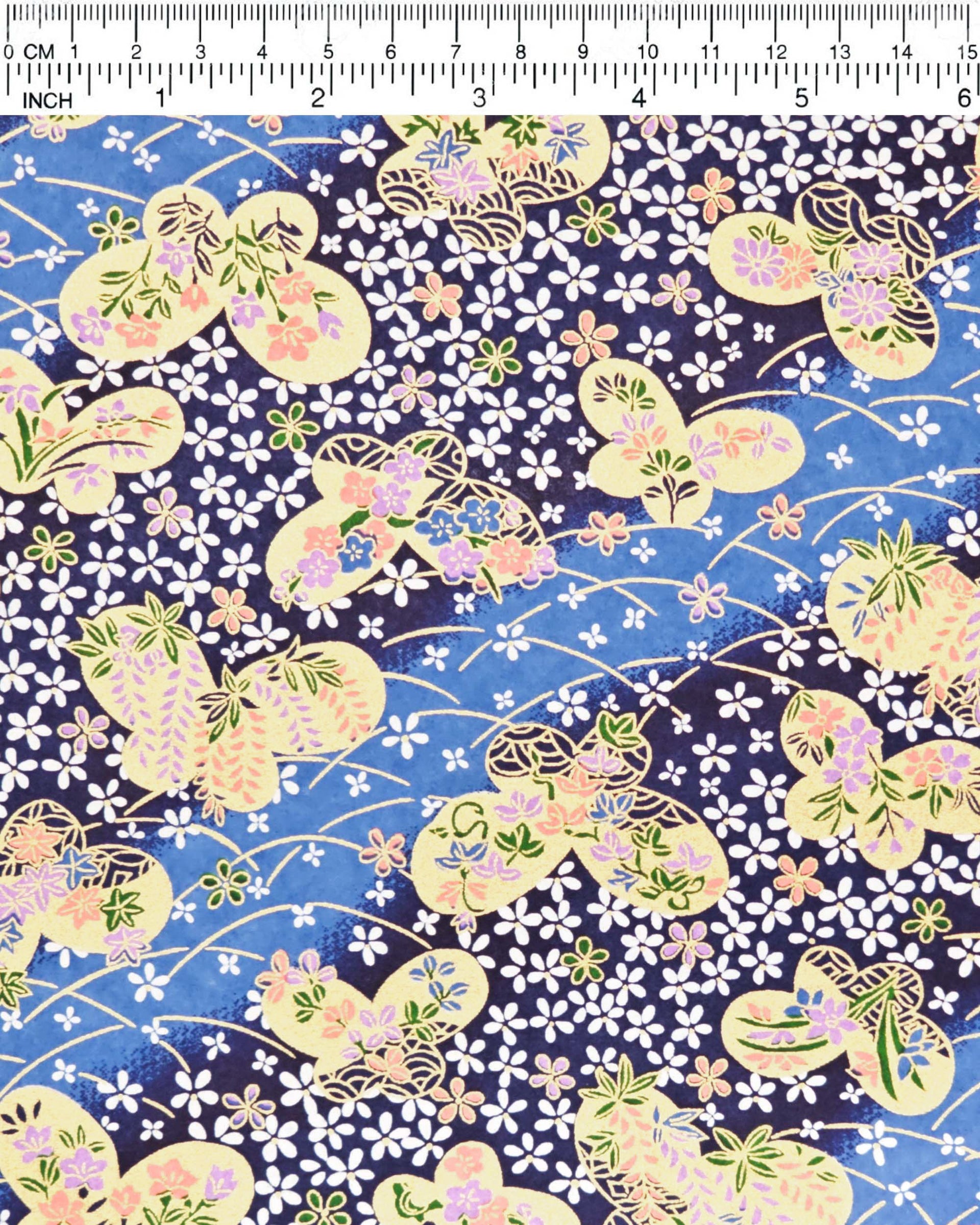 Japanese chiyogami paper featuring metallic gold butterflies with purple and pink flowers on a blue background covered with small white flowers, shown next to a ruler for size reference.