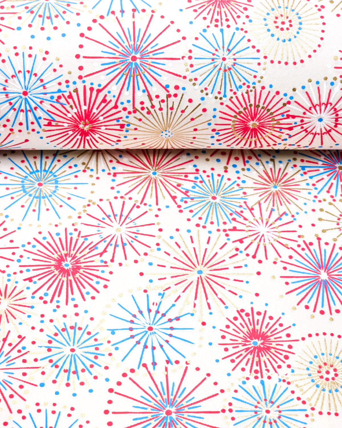 Japanese chiyogami paper featuring colorful red, blue, pink, and white exploding fireworks on a white background with metallic gold details, shown rolled to highlight the design.