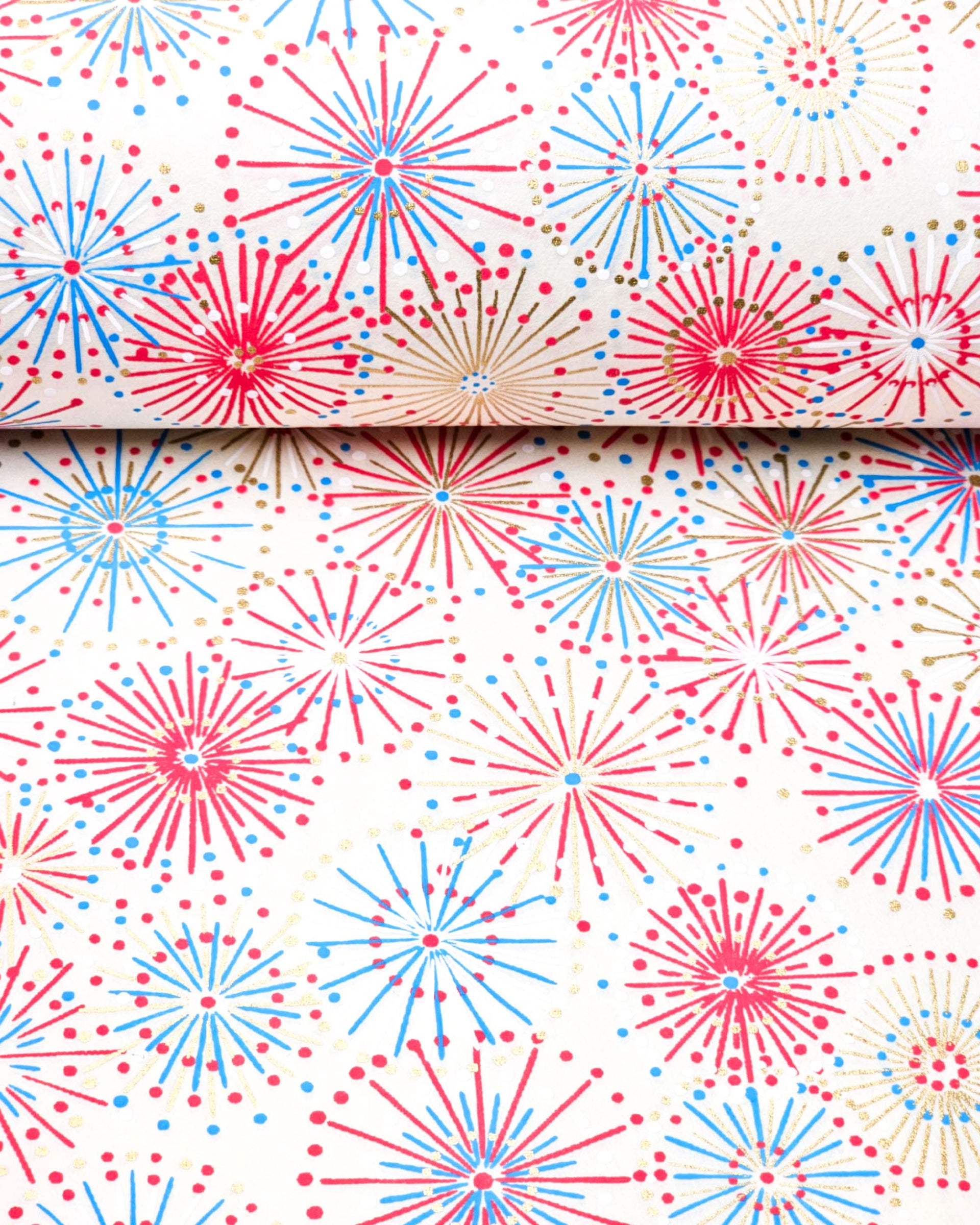 Japanese chiyogami paper featuring colorful red, blue, pink, and white exploding fireworks on a white background with metallic gold details, shown rolled to highlight the design.