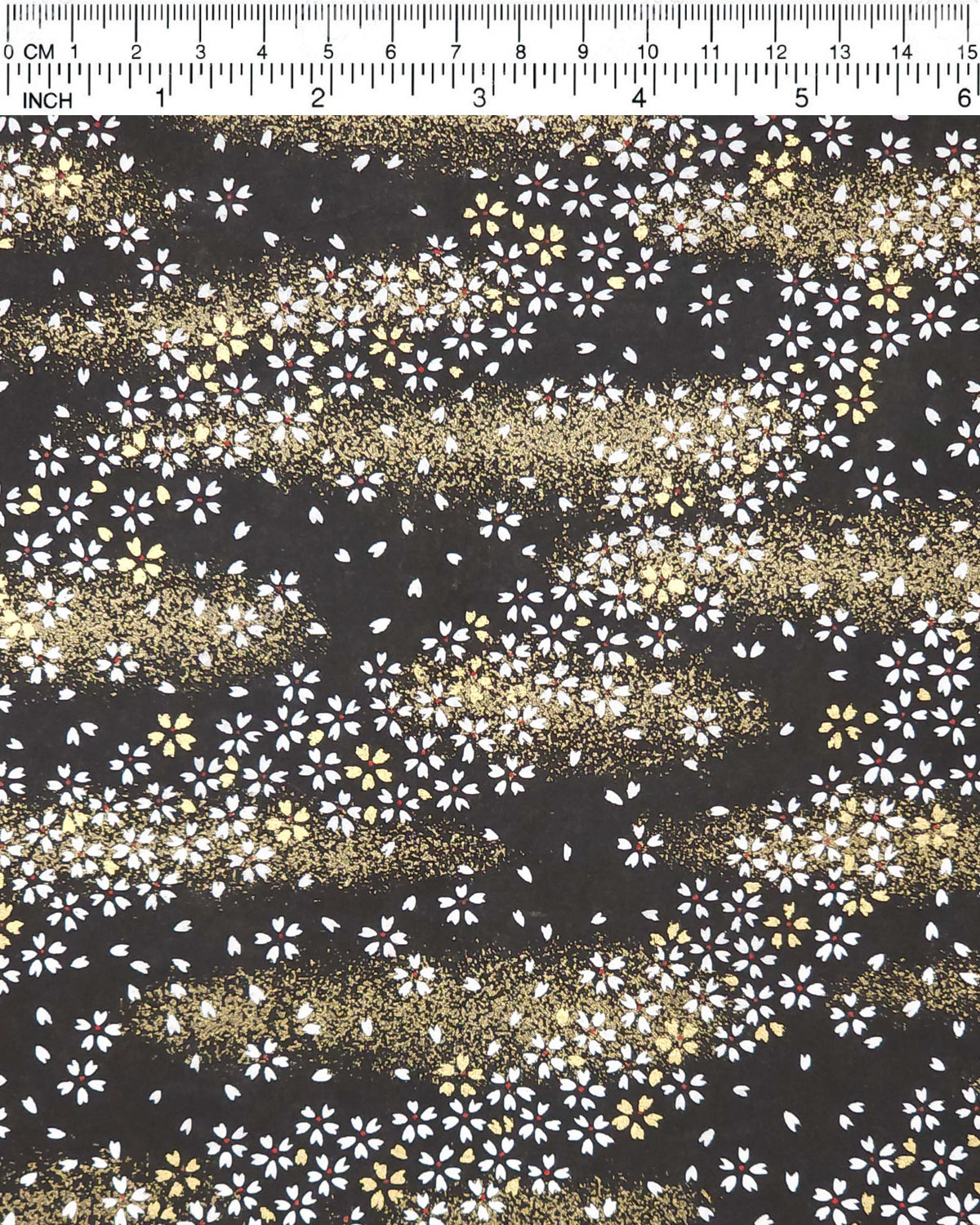 Japanese chiyogami paper featuring small white and gold cherry blossoms on a black background with metallic gold details, shown next to a ruler for size reference.