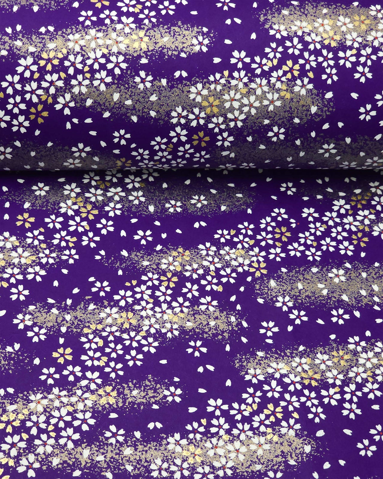 Japanese chiyogami paper featuring small white and gold cherry blossoms on a dark blue background with metallic gold details, shown rolled to highlight the design.
