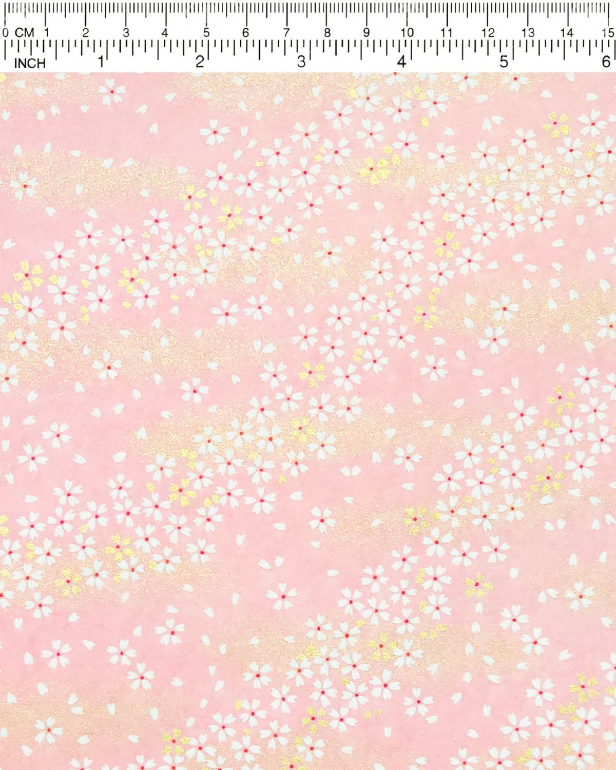 Japanese chiyogami paper featuring small white and gold cherry blossoms on a pink background with metallic gold details, shown next to a ruler for size reference.