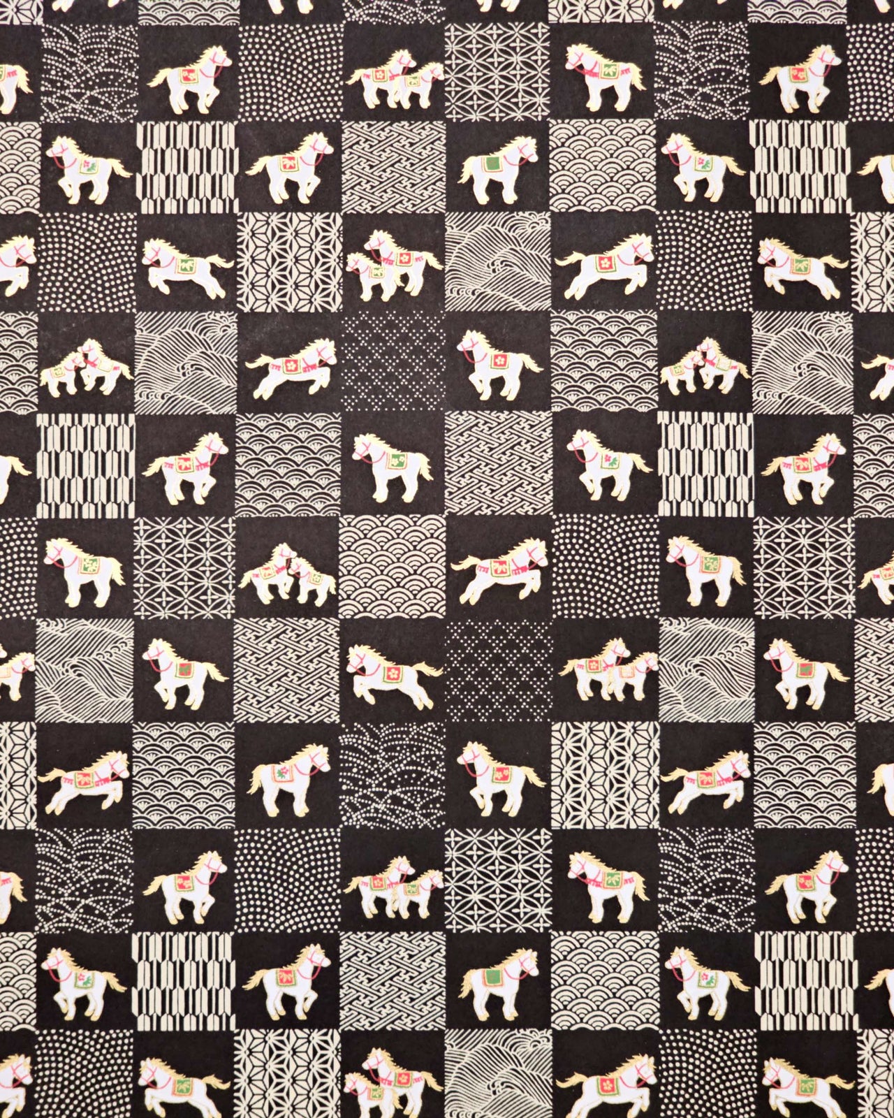 Japanese chiyogami paper featuring white horses with gold manes wearing red and green saddles and bridles, alternating with assorted off-white traditional Japanese patterns in an ichimatsu (checker) design on a black background.