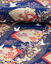 Japanese chiyogami paper featuring white flying cranes, gold and red folding fans, and assorted red, purple, white, and yellow flowers on a dark blue background with metallic gold wave and geometric details, shown rolled to highlight the design.