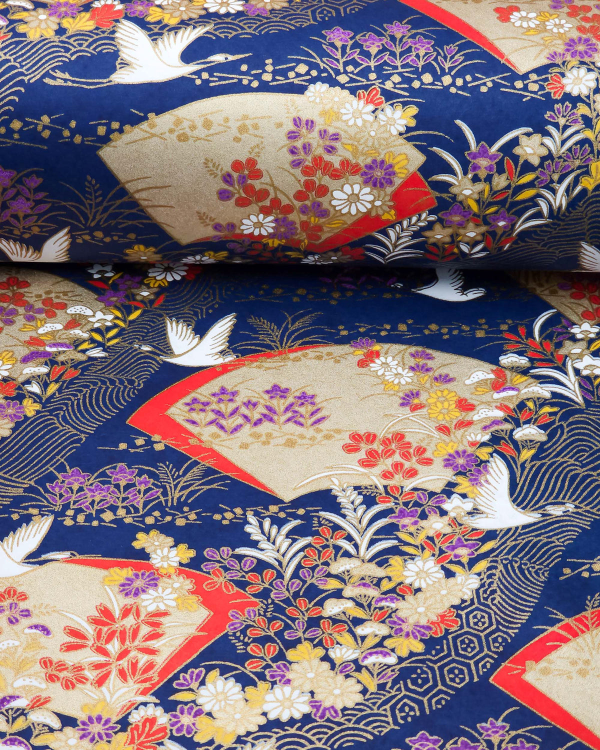 Japanese chiyogami paper featuring white flying cranes, gold and red folding fans, and assorted red, purple, white, and yellow flowers on a dark blue background with metallic gold wave and geometric details, shown rolled to highlight the design.