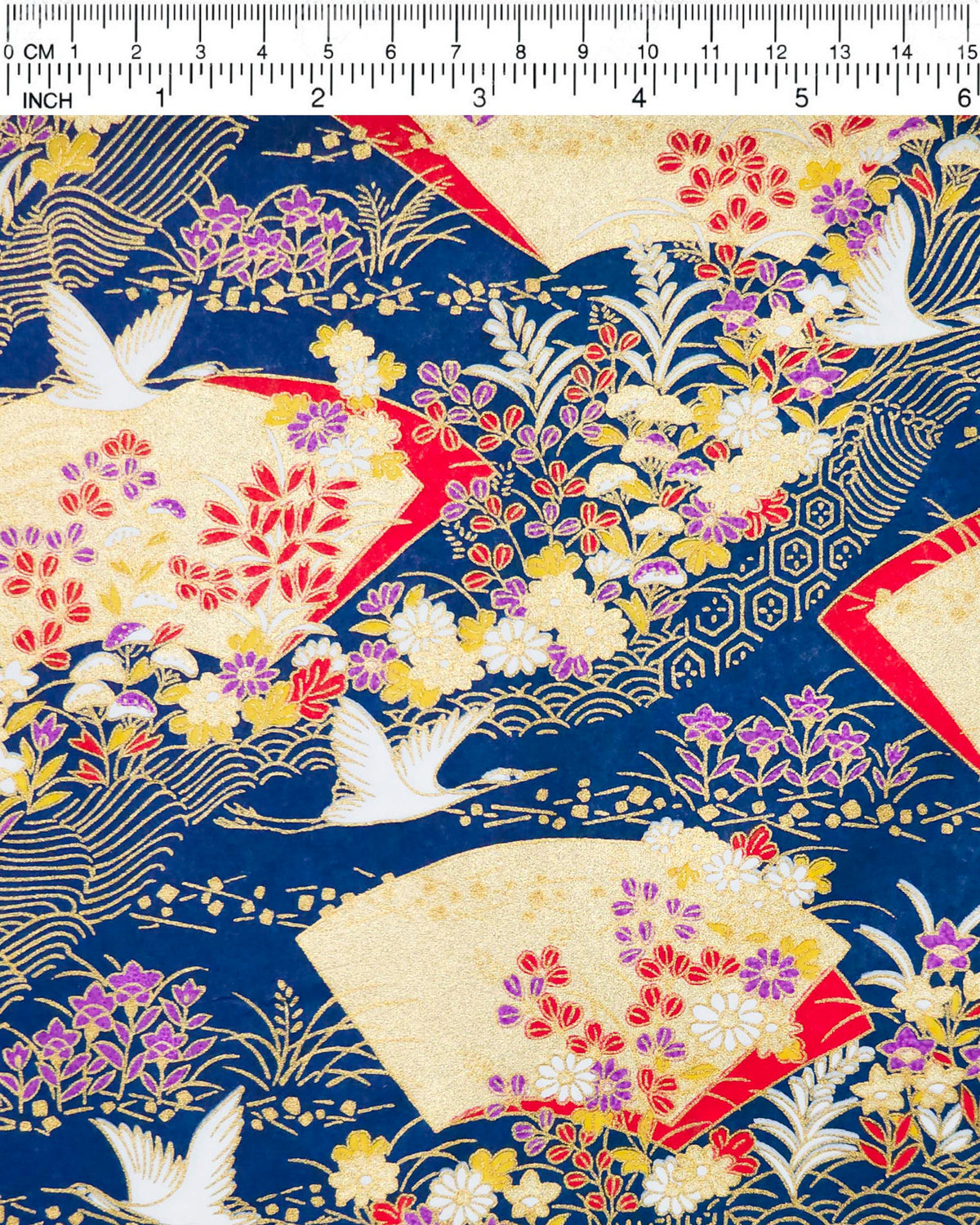Japanese chiyogami paper featuring white flying cranes, gold and red folding fans, and assorted red, purple, white, and yellow flowers on a dark blue background with metallic gold wave and geometric details, shown next to a ruler for size reference.
