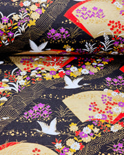 Japanese chiyogami paper featuring white flying cranes, gold and red folding fans, and assorted red, purple, white, and yellow flowers on a black background with metallic gold wave and geometric details, shown rolled to highlight the design.