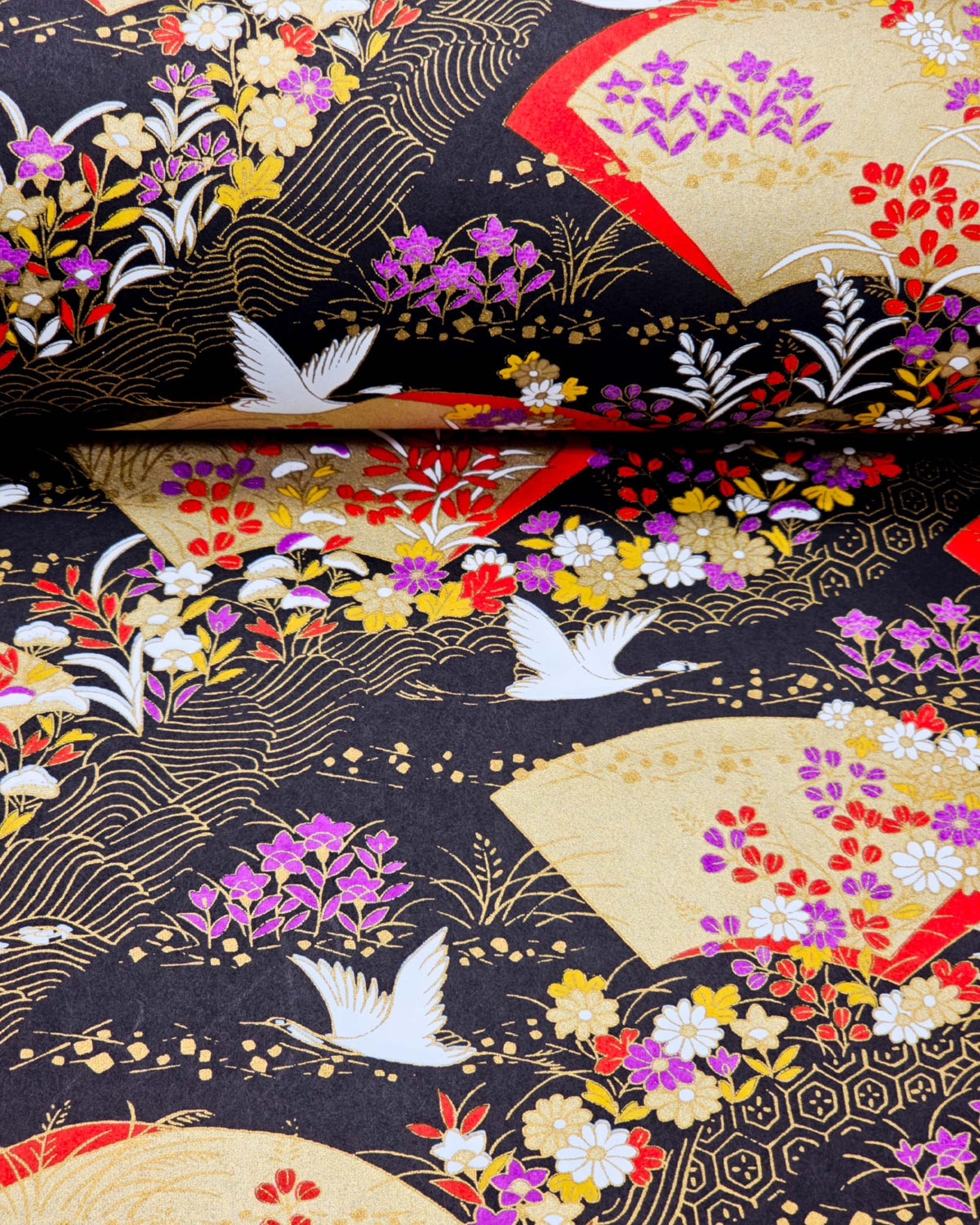 Japanese chiyogami paper featuring white flying cranes, gold and red folding fans, and assorted red, purple, white, and yellow flowers on a black background with metallic gold wave and geometric details, shown rolled to highlight the design.