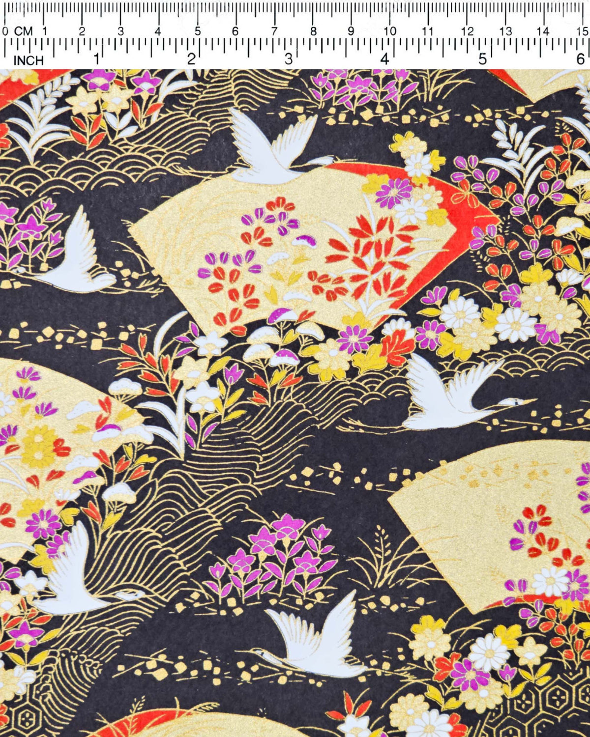 Japanese chiyogami paper featuring white flying cranes, gold and red folding fans, and assorted red, purple, white, and yellow flowers on a black background with metallic gold wave and geometric details, shown next to a ruler for size reference.