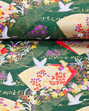 Japanese chiyogami paper featuring white flying cranes, gold and red folding fans, and assorted red, purple, white, and yellow flowers on a green background with metallic gold wave and geometric details, shown rolled to highlight the design.