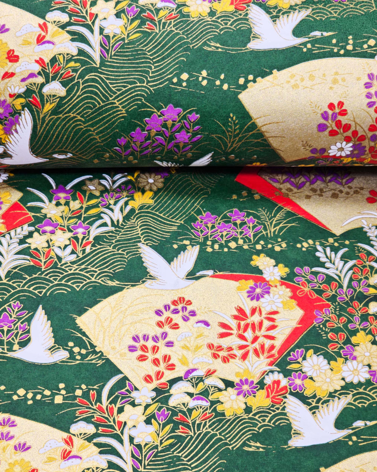 Japanese chiyogami paper featuring white flying cranes, gold and red folding fans, and assorted red, purple, white, and yellow flowers on a green background with metallic gold wave and geometric details, shown rolled to highlight the design.