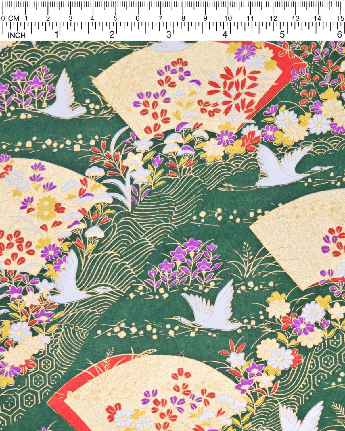 Japanese chiyogami paper featuring white flying cranes, gold and red folding fans, and assorted red, purple, white, and yellow flowers on a green background with metallic gold wave and geometric details, shown next to a ruler for size reference.