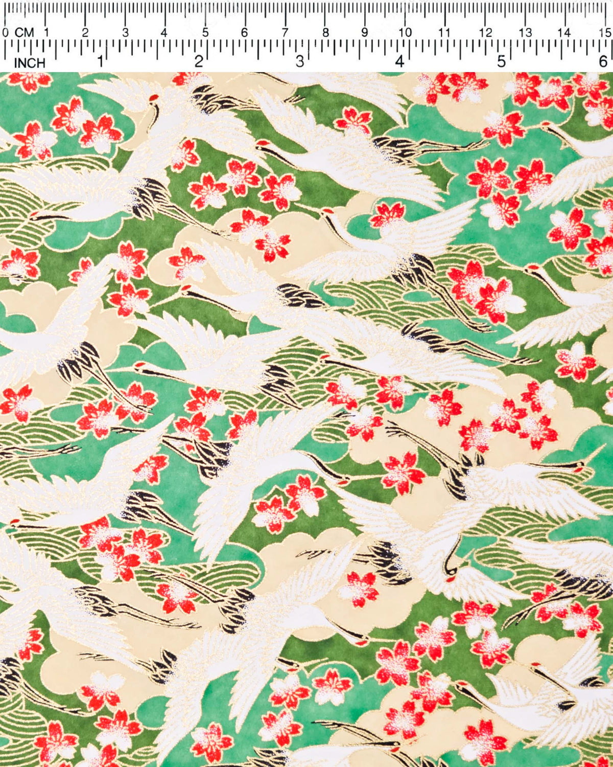 Japanese chiyogami paper featuring white flying cranes and red and white cherry blossoms among beige and green clouds and green waves with metallic gold details, shown next to a ruler for size reference.