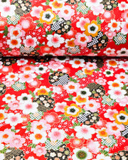 Japanese chiyogami paper featuring pink, white, and orange cherry blossoms with green leaves and black yukiwa (snowflake) on a red background with metallic gold details, shown rolled to highlight the design.