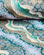 Japanese chiyogami paper featuring pink and white cherry blossoms with assorted traditional Japanese floral and geometric patterns on teal blue background with metallic gold and silver details, shown rolled to highlight the design.